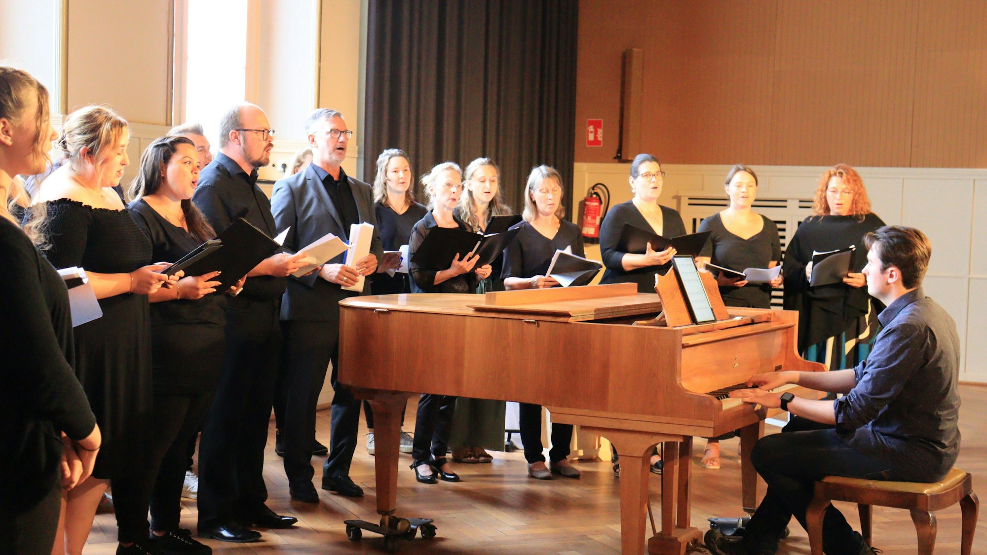 Sängerinnen und Sänger, alle in Schwarz gekleidet, haben sich um einen Flügel gruppiert, auf dem ein Pianist spielt.