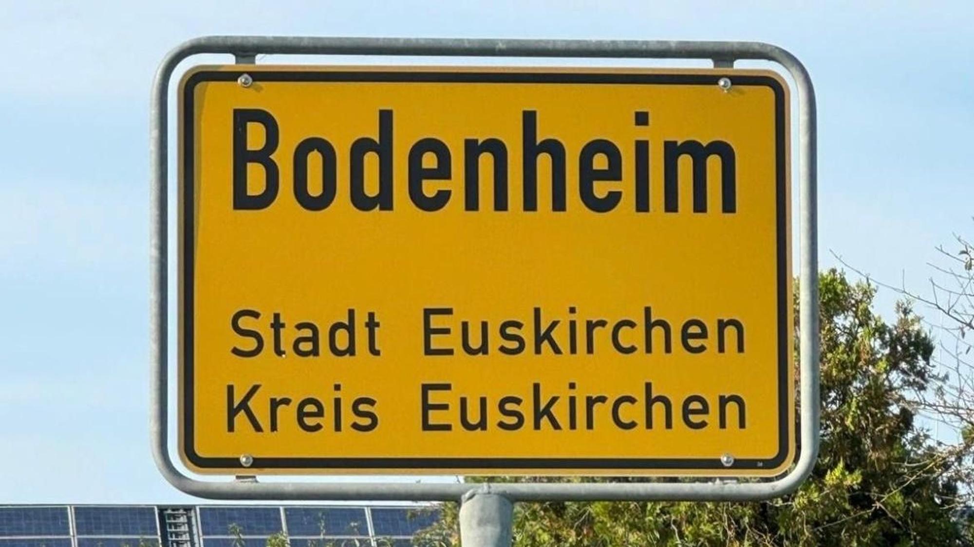 Ein gelbes Ortsschild mit der Aufschrift: „Bodenheim, Stadt Euskirchen, Kreis Euskirchen“ steht vor grünen Büschen und blauem Himmel.