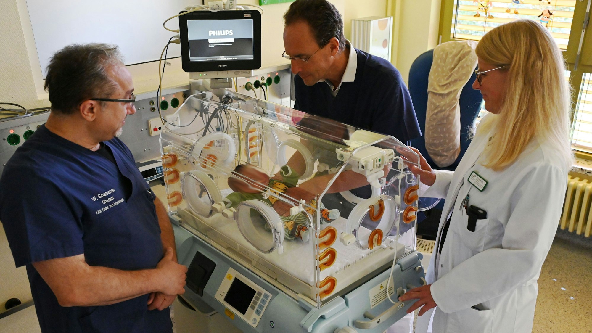 Zwei Männer in blauen Shirts und eine Frau in weißem Arztkittel, Wael Shabanah, Dr. Herbert Schade und Dr. Tatjana Klug betrachten ein neugeborenes Kind auf der Kinderintensivstation im Kreiskrankenhaus Mechernich.