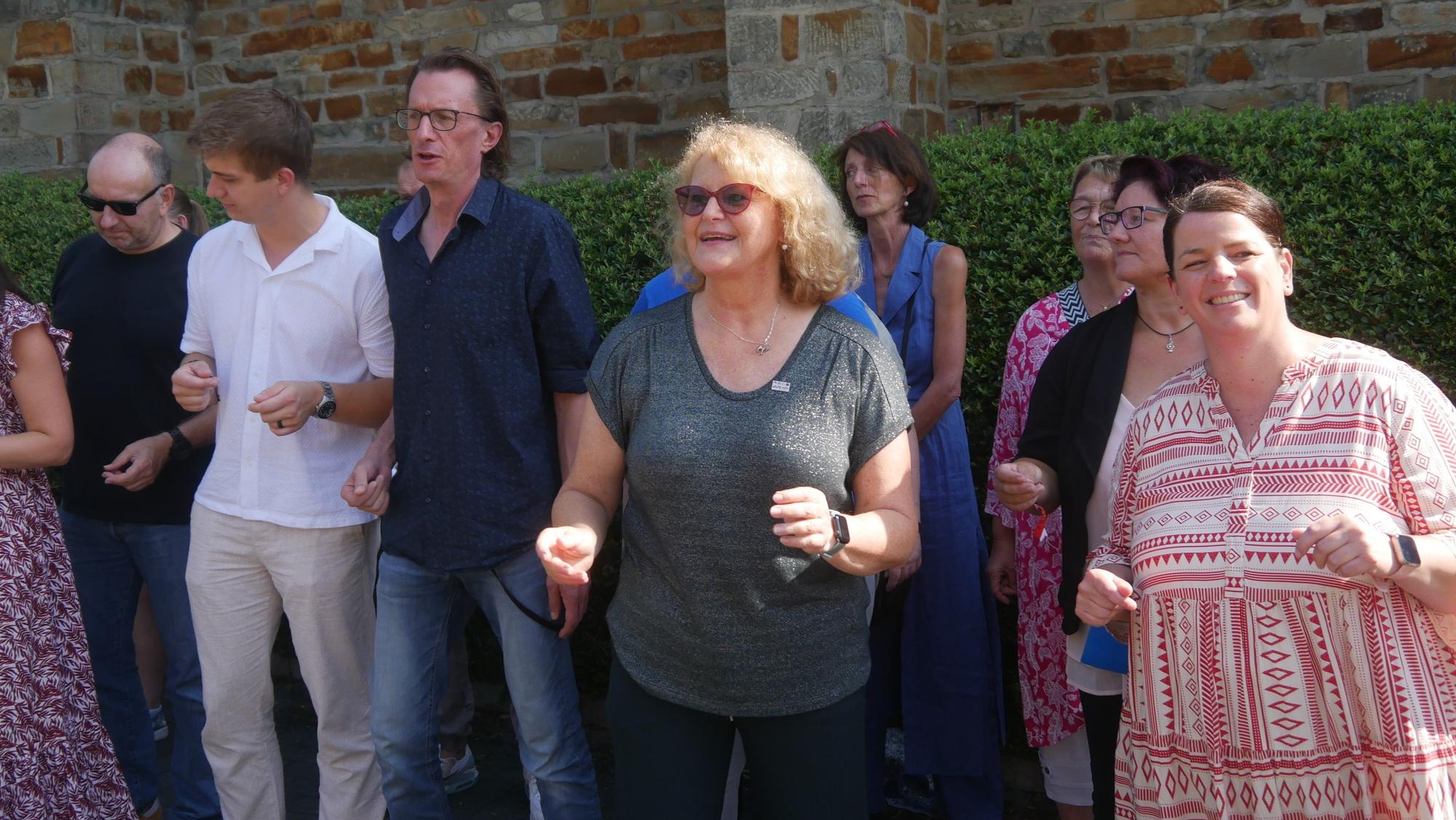 Eine blonde Frau mit Brille in einer Reihe von tanzenden und singenden Menschen.