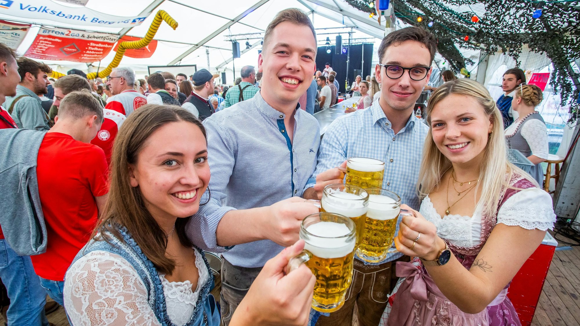 Zwei junge Frauen und zwei junge Männer in Tracht prosten sich mit Maßkrügen zu.