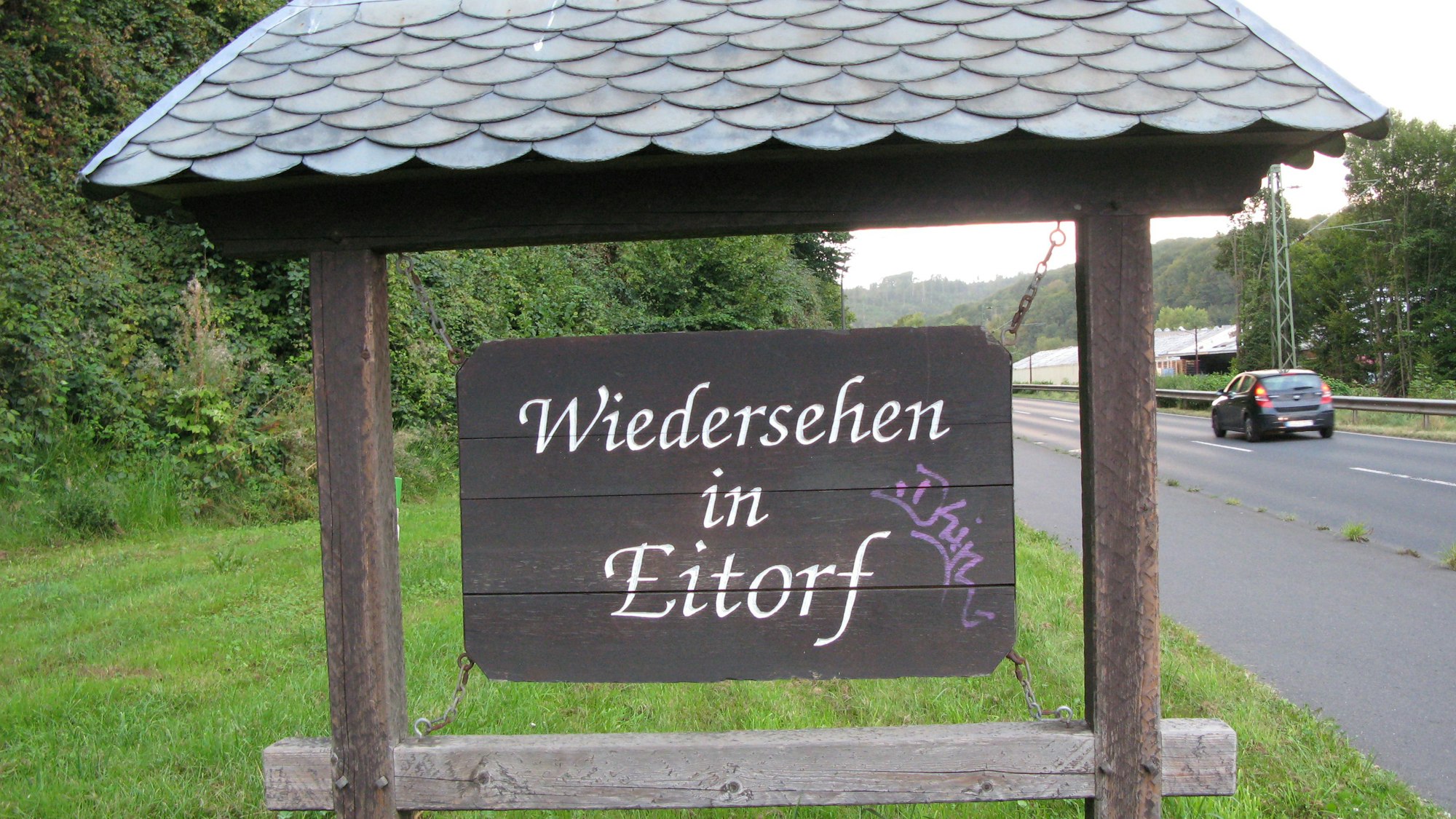 Ein altes, 70 mal 110 Zentimeter großes Holzschild in einem überdachten Balkenrahmen mit der Inschrift „Wiedersehen in Eitorf“ an einer Straße.