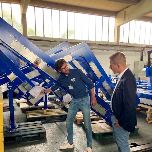 Ein Mann im Hemd zeigt mit einem Bauplan in der Hand auf ein Winkelförderband in einer Halle, während ihm ein Mann im Anzug zuschaut.