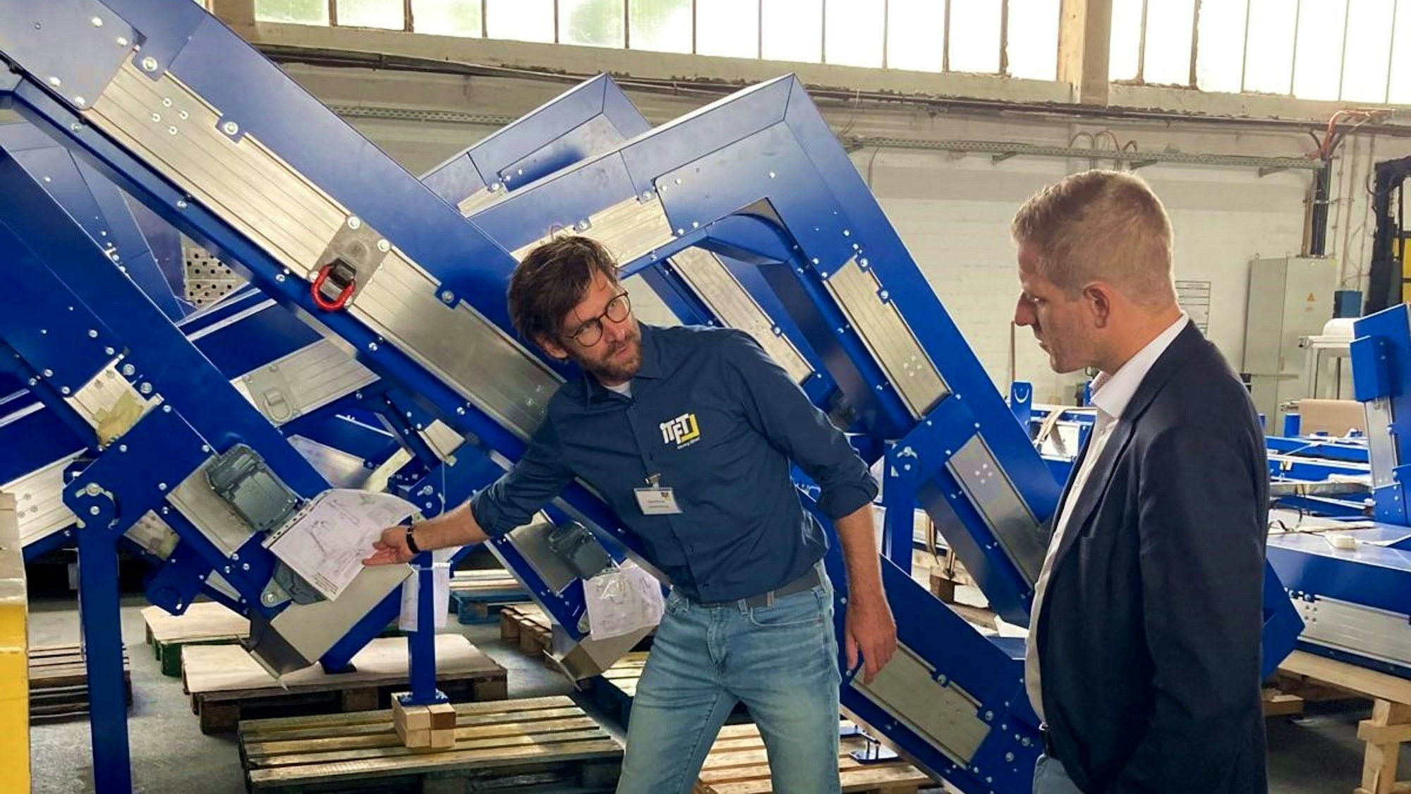 Ein Mann im Hemd zeigt mit einem Bauplan in der Hand auf ein Winkelförderband in einer Halle, während ihm ein Mann im Anzug zuschaut.
