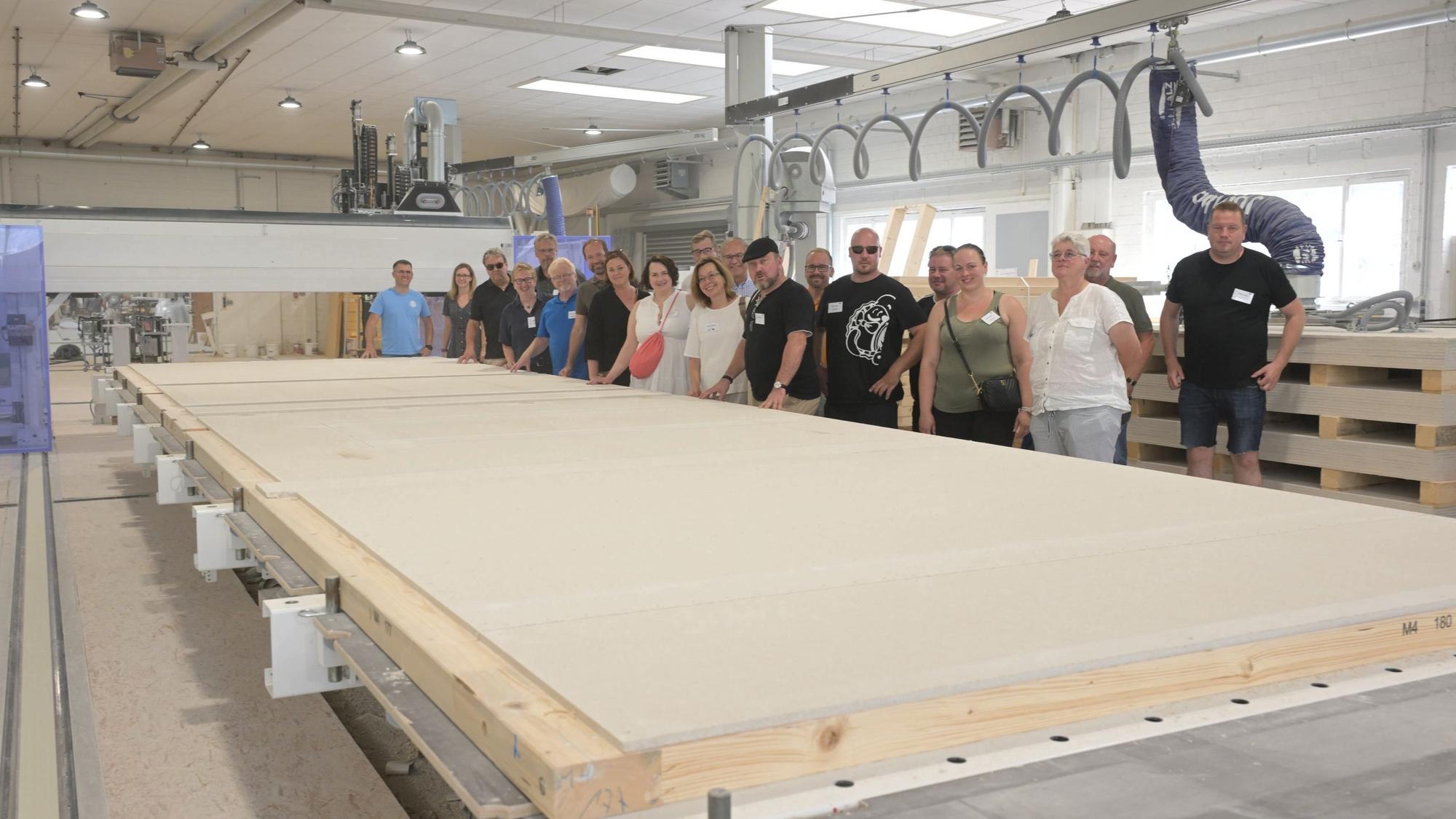 Eine Gruppe von Menschen steht in einer großen Fertigungshalle. Vor ihnen liegt ein große Holzplatte, die einmal eine Hauswand werden soll.