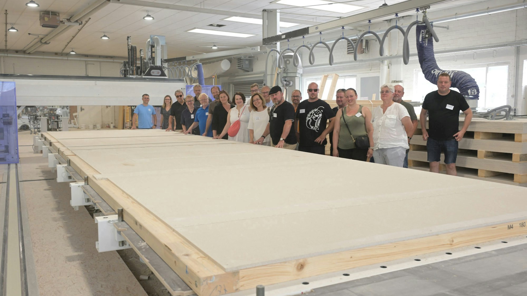 Eine Gruppe von Menschen steht in einer großen Fertigungshalle. Vor ihnen liegt ein große Holzplatte, die einmal eine Hauswand werden soll.