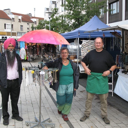 Zwei Markthändler und eine Markthändlerin stehen an einem Schmuckstand auf dem Marktplatz.