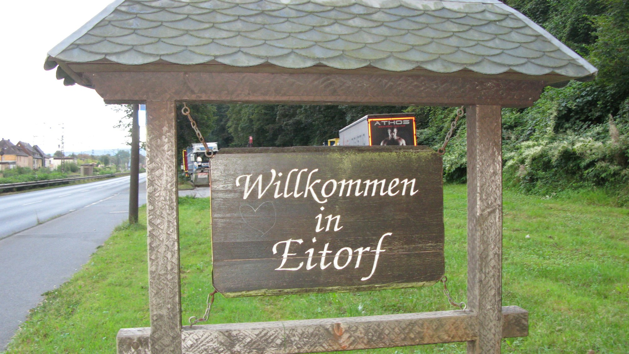 Ein altes, 70 mal 110 Zentimeter großes Holzschild in einem überdachten Balkenrahmen mit der Inschrift „Willkommen in Eitorf“ an einer Straße.