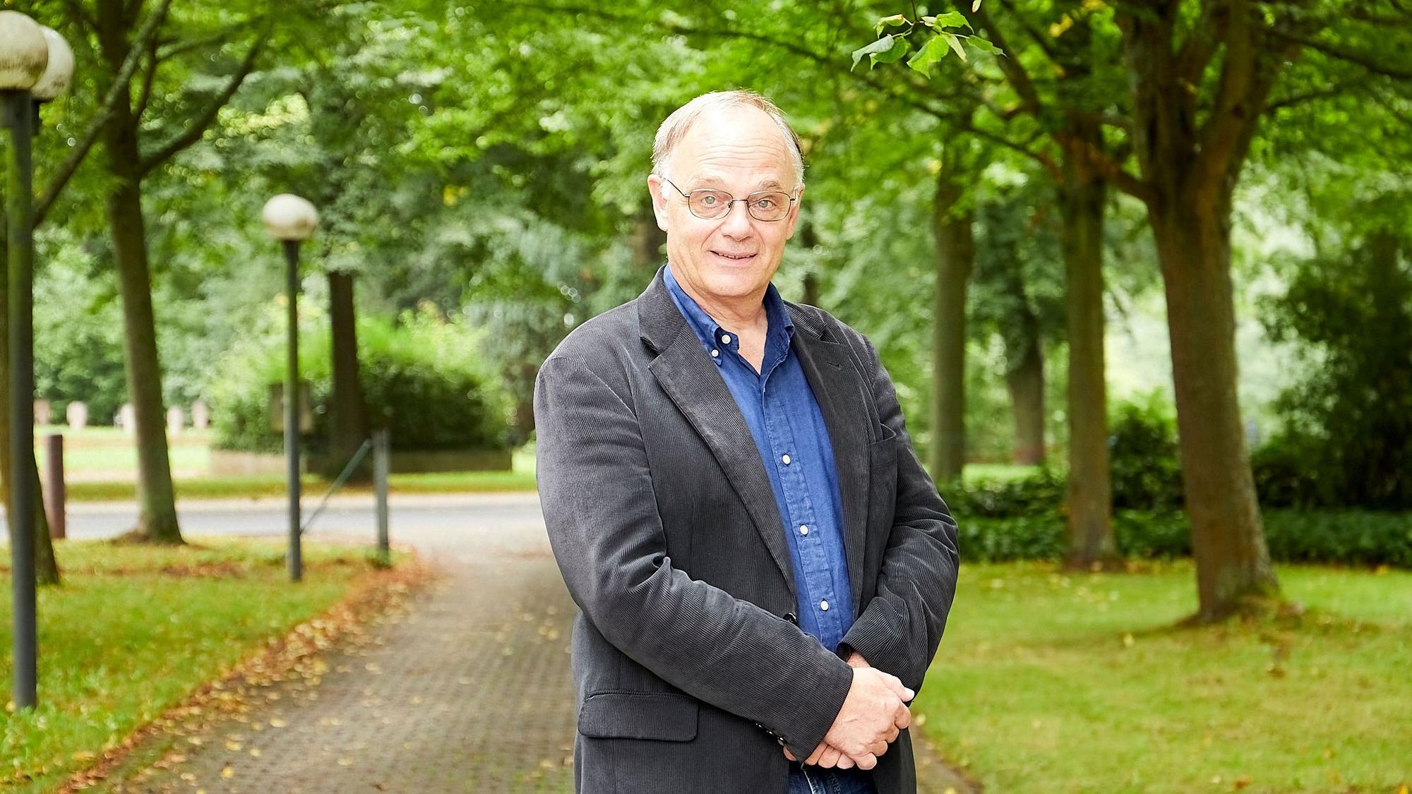 Ein Herr mit Brille legt in einem Park die Hände vor dem Bauch übereinander.