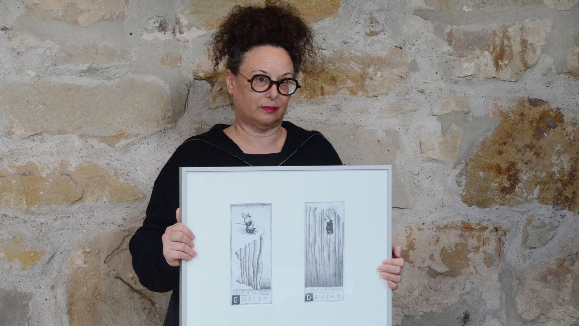 Eine Frau im schwarzen Kleid steht vor einer grob gemauerten Wand. Sie hält einen Bilderrahmen mit zwei Schwarz-Weiß-Zeichnungen in den Händen.
