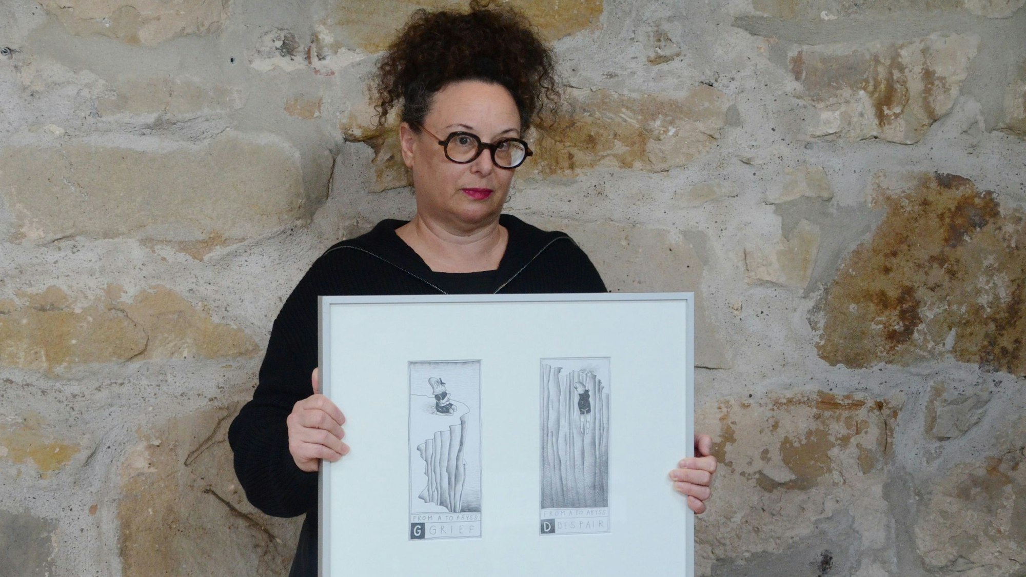 Eine Frau im schwarzen Kleid steht vor einer grob gemauerten Wand. Sie hält einen Bilderrahmen mit zwei Schwarz-Weiß-Zeichnungen in den Händen.