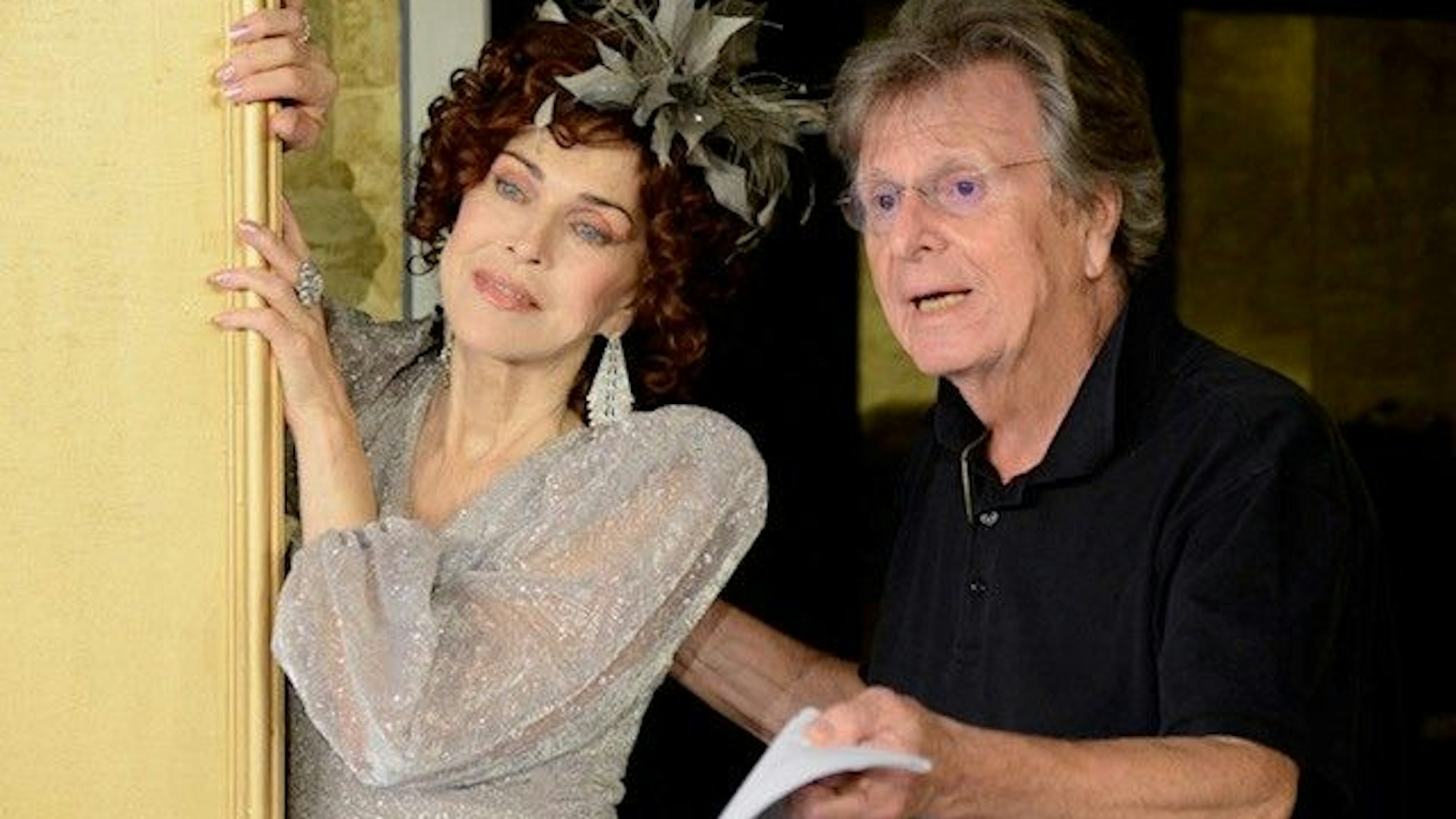 Eine Frau im Kostüm einer Operndiva und ein Mann mit Brille und im schwarzen Polohemd bei einer Probe auf der Theaterbühne.