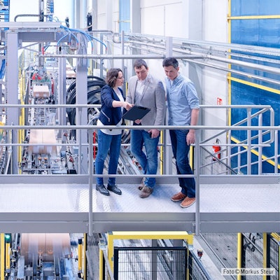 Eine Frau und zwei Männer stehen in einer Fabrikhalle.