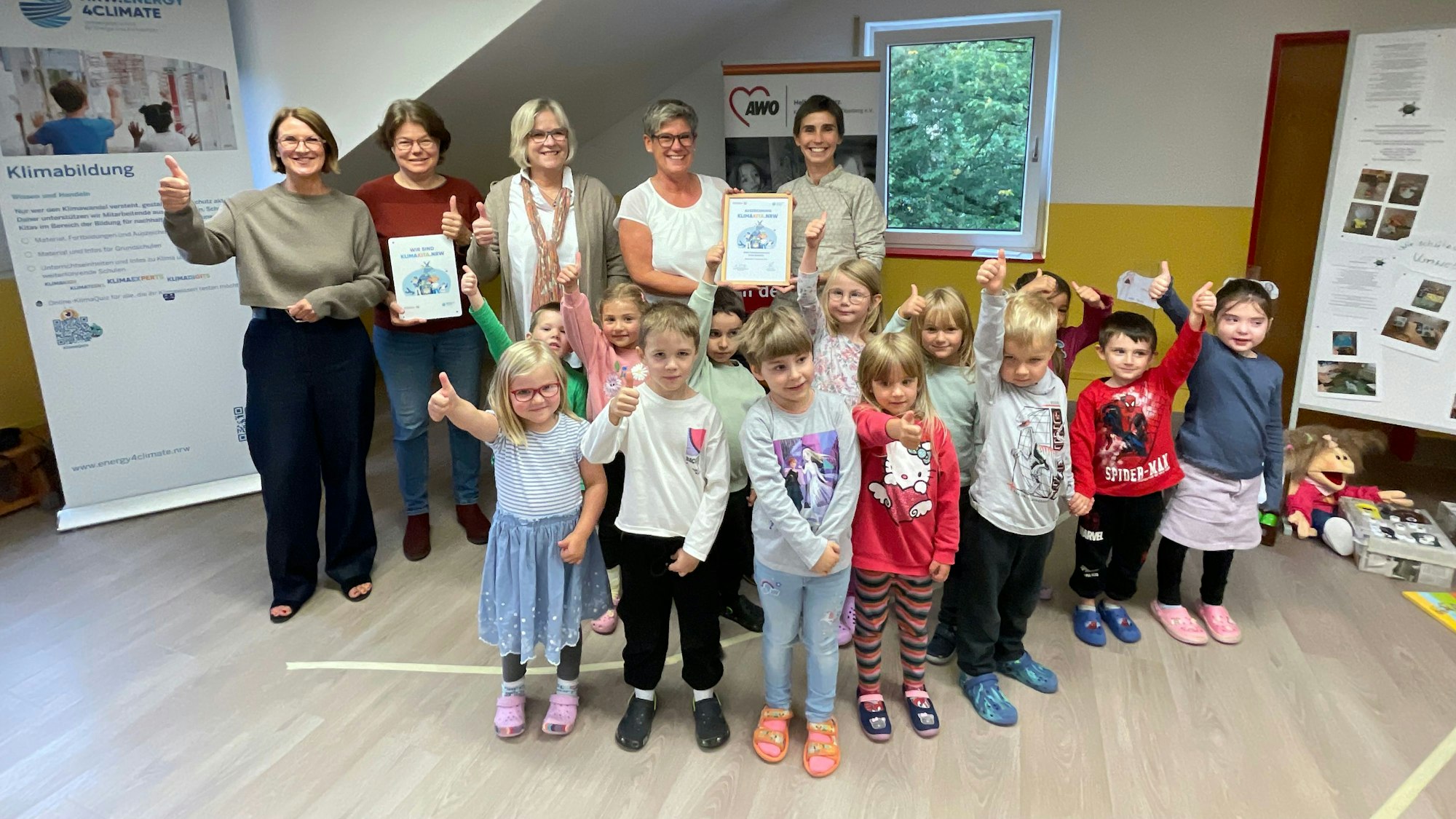 Eine Gruppe Kinder im Kindergartenalter steht gemeinsam mit mehreren Erwachsenen in einem bunten Zimmer und zeigen die „Daumen hoch“ Geste.