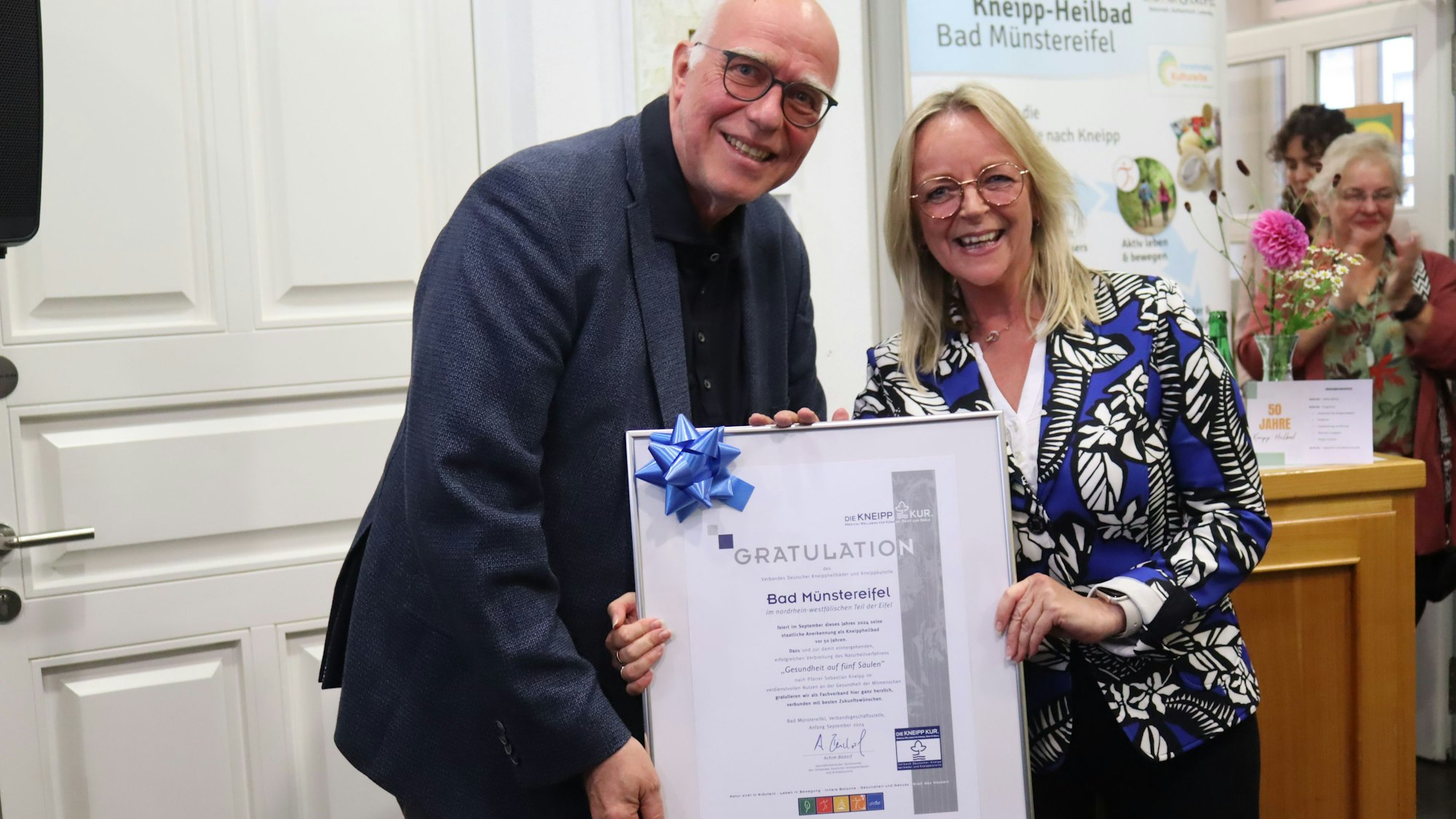 Ein Mann und eine Frau halten eine Urkunde in die Kamera. Beide freuen sich. Der Mann ist größer und muss sich hinabbeugen.