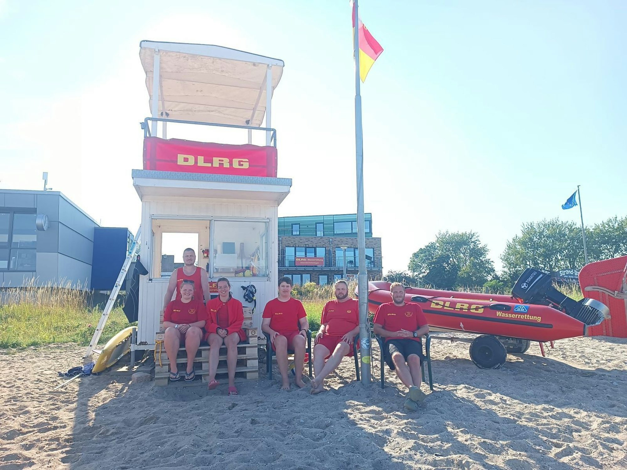 Gruppenbild von sechs Rettungsschwimmern vor einem Wachturm, rechts im Bild ist ein Schlauchboot der Wasserrettung zu sehen.