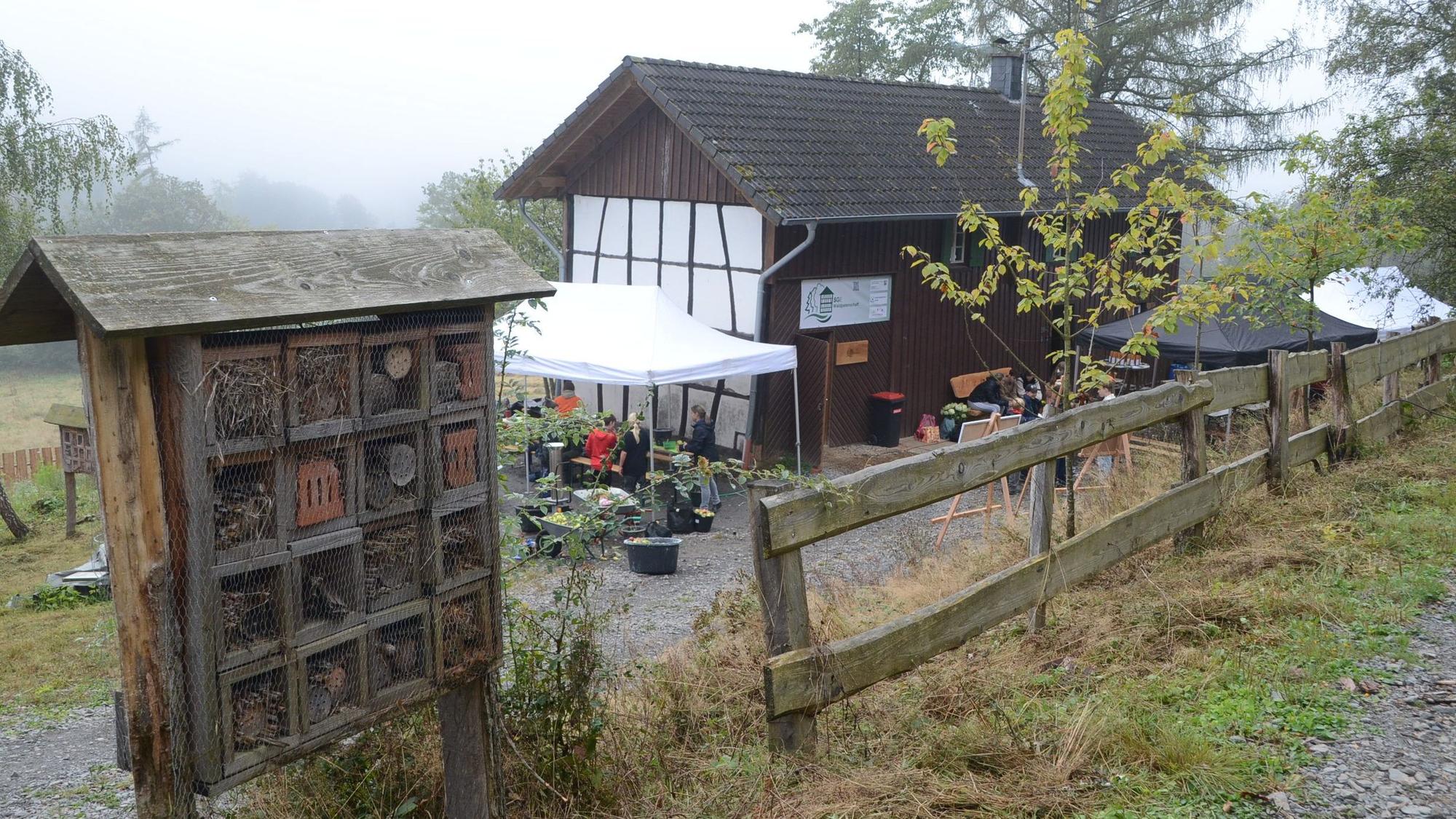 Ein Holzhaus mit Fachwerk an der Giebelwand. Im Vordergrund steht ein großes „Insektenhotel“.