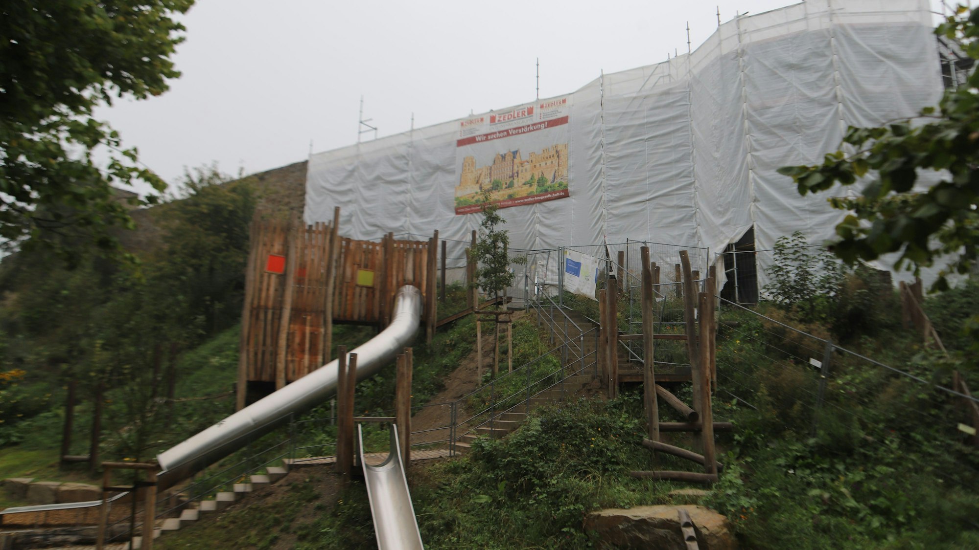 Hinter einem Spielplatz ist eine Stadtmauer von einer Plane verhängt.