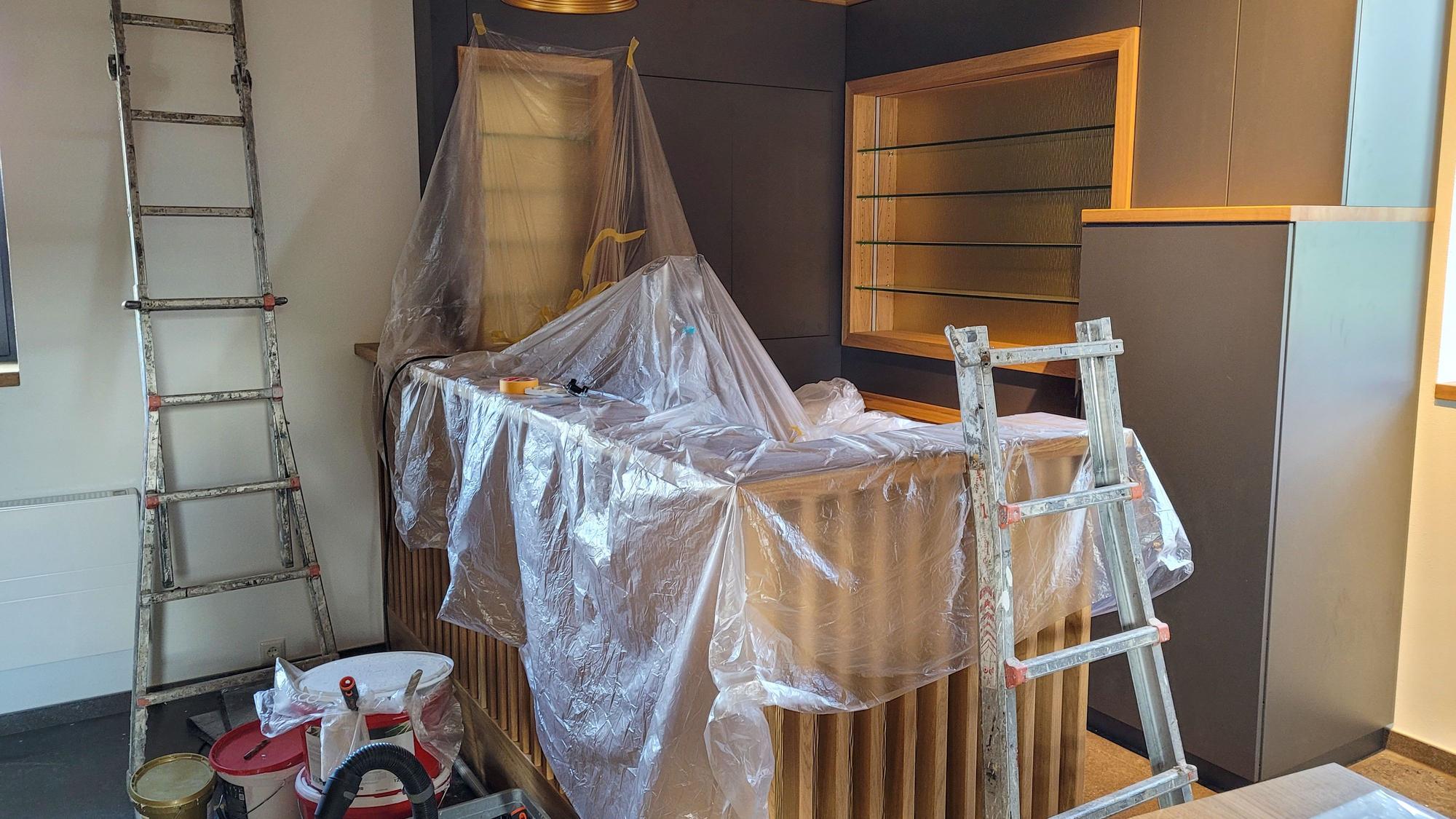 Unter durchsichtiger Malerfolie ist eine Holztheke zu erkennen, davor stehen Farbeimer und Pinsel, an der Wand lehnt eine Leiter.