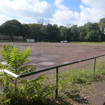 Im Vordergrund ist das Geländer des Aschenplatzes zu sehen. Auf dem Platz parkt ein Auto.