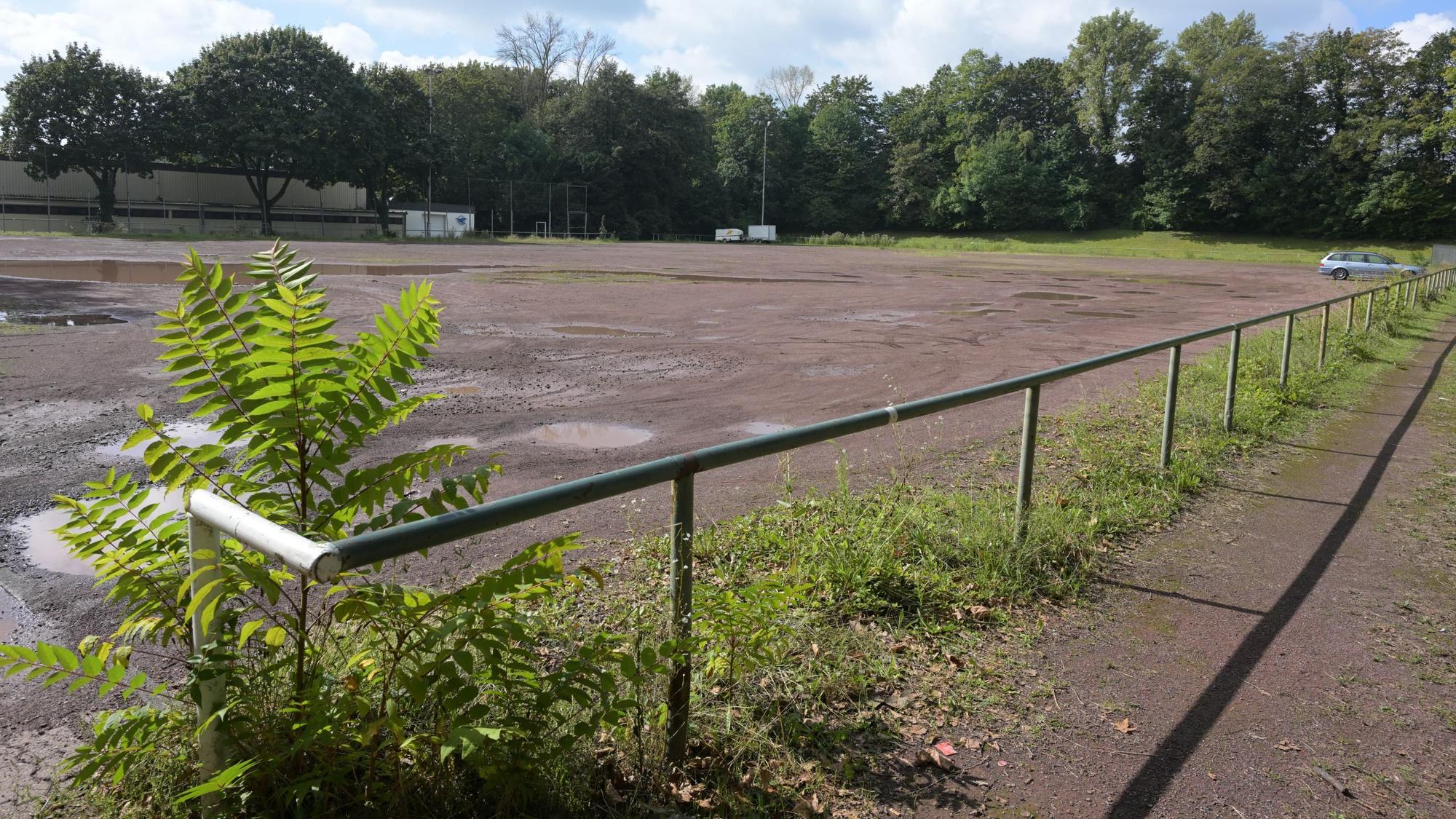 Im Vordergrund ist das Geländer des Aschenplatzes zu sehen. Auf dem Platz parkt ein Auto.