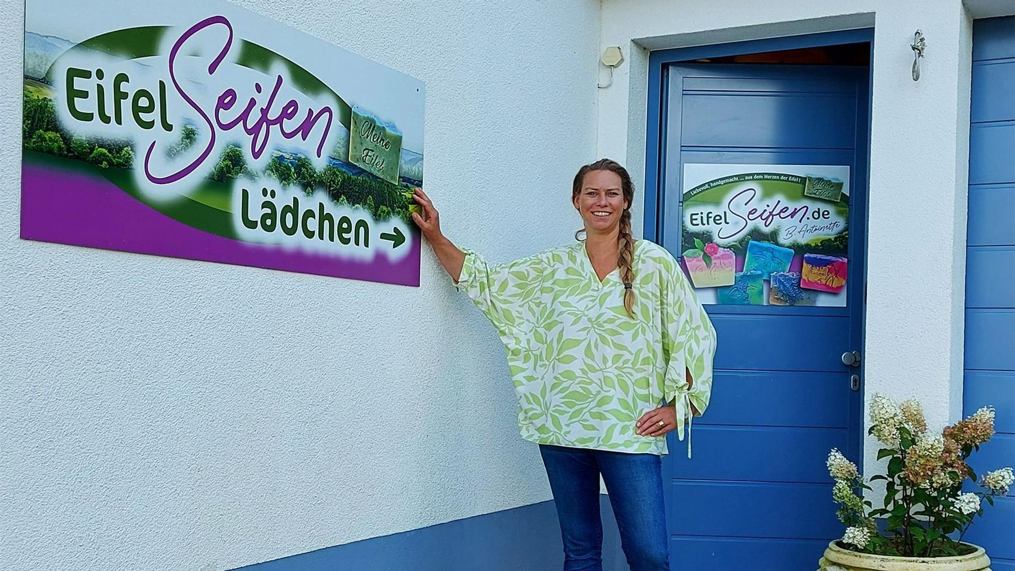 Eine Frau steht an einem Wohnhaus in Blankenheim mit einer blauen Tür und deutet auf ein Schild mit der Aufschrift „Eifelseifen Lädchen“ hin.