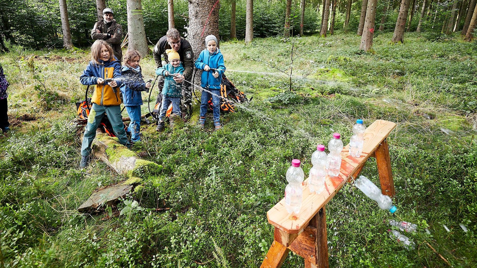 Vier Kinder stehen im Wald und spitzen Wasser aus Löschrucksäcken auf leere Plastikflaschen, um sie in einem Spiel von einem kleinen Tisch zu schießen.