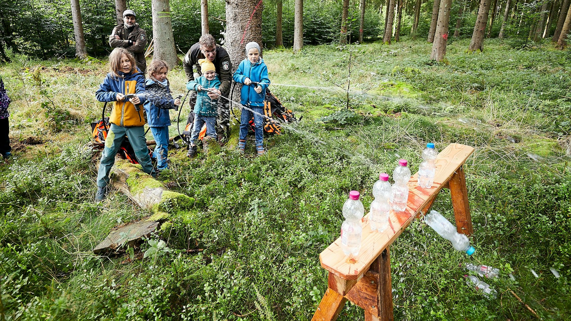Vier Kinder stehen im Wald und spitzen Wasser aus Löschrucksäcken auf leere Plastikflaschen, um sie in einem Spiel von einem kleinen Tisch zu schießen.