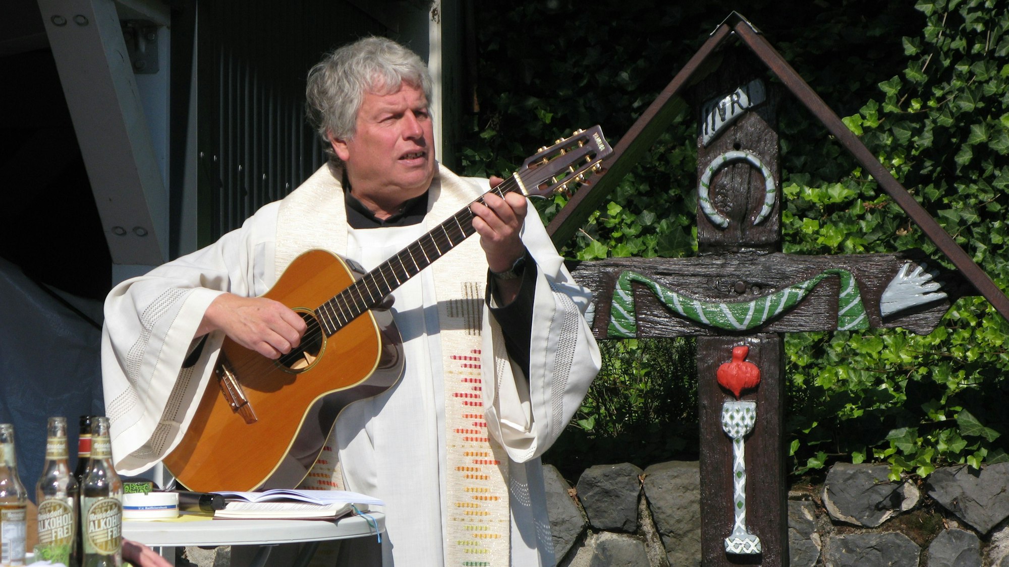 Ein Pfarrer spielt Gitarre und singt. Neben ihm steht das restaurierte Holzkreuz.