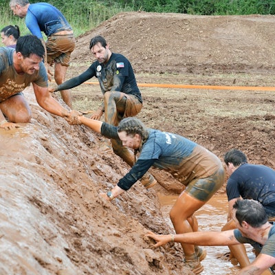 Mehrere Teilnehmerinnen und Teilnehmer überqueren einen verschlammten Damm. Ein Mann reicht einer Frau die Hand, um ihr zu helfen.