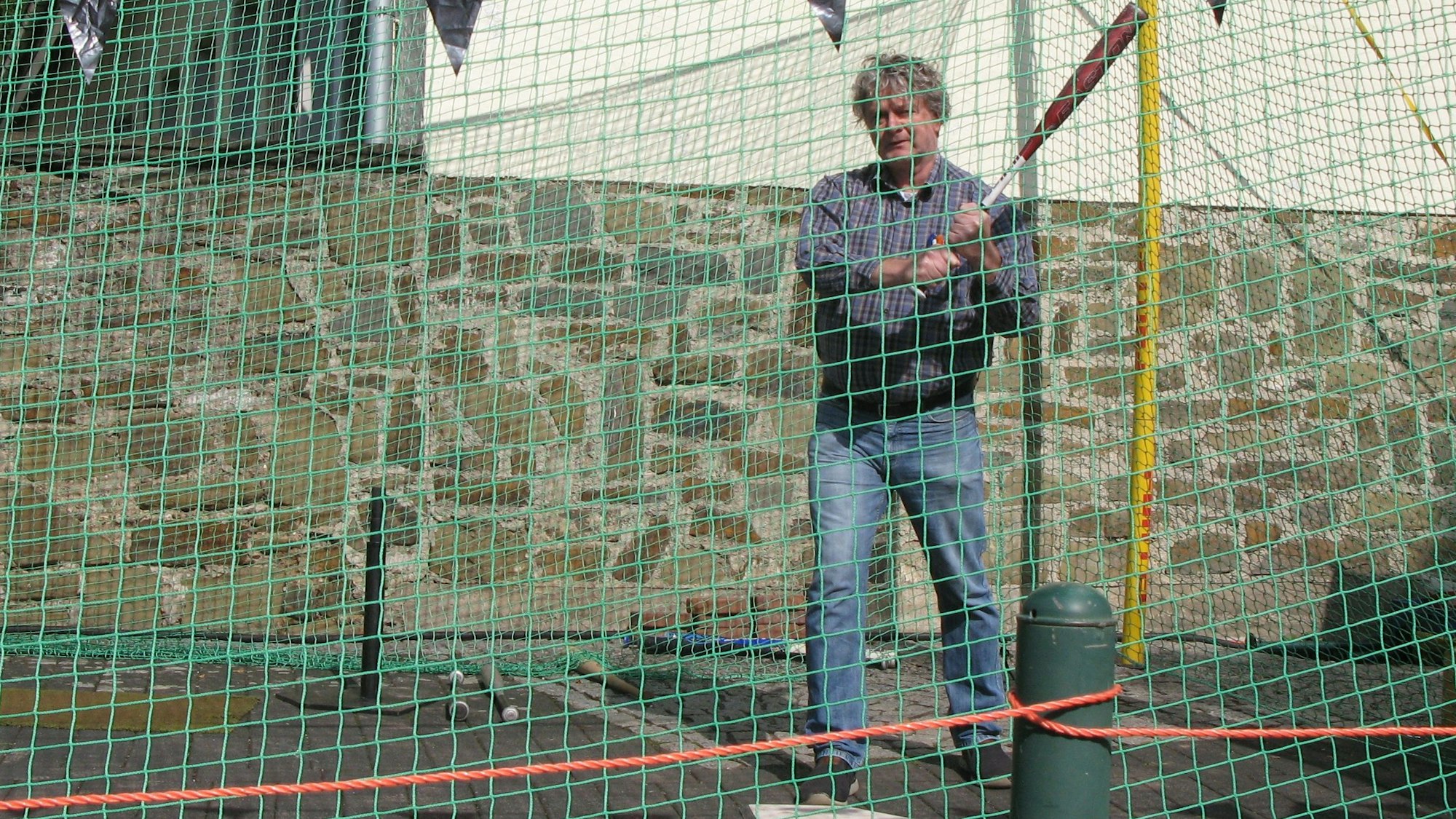 Mann holt mit einem Baseballschläger hinter einem Fangnetz zum Schlag aus.