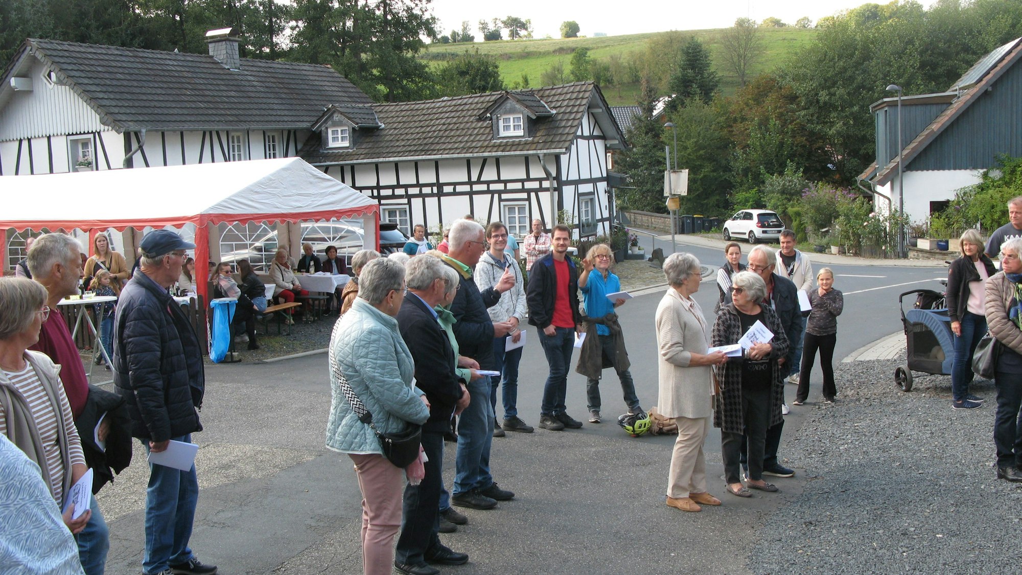 Etwa zwei Dutzend Menschen stehen an einer Straße, im Hintergrund sitzen weitere in einem Partyzelt.