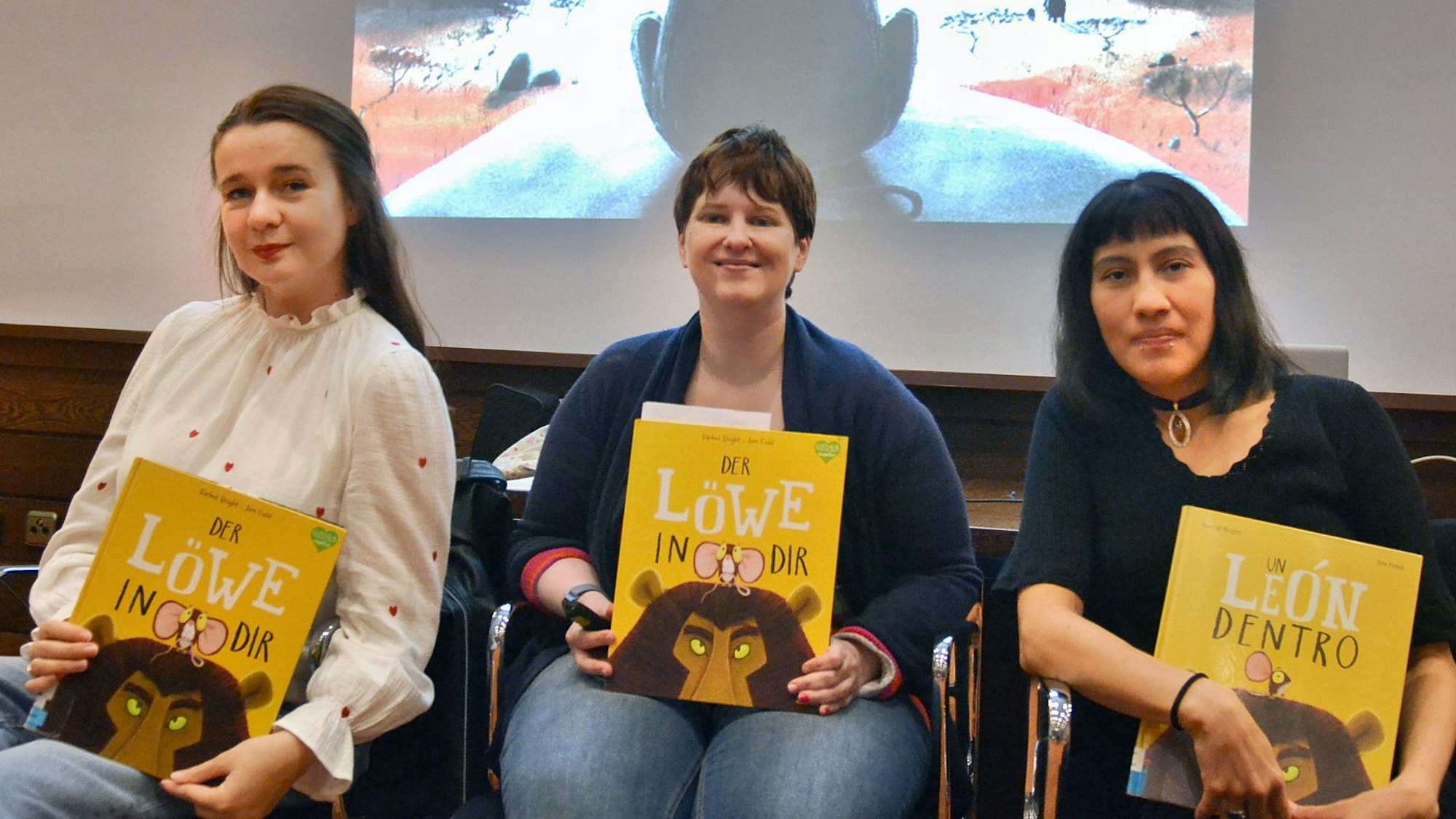 Drei Frauen sitzen auf Stühlen nebeneinander, sie halten jeweils ein Buch fest.