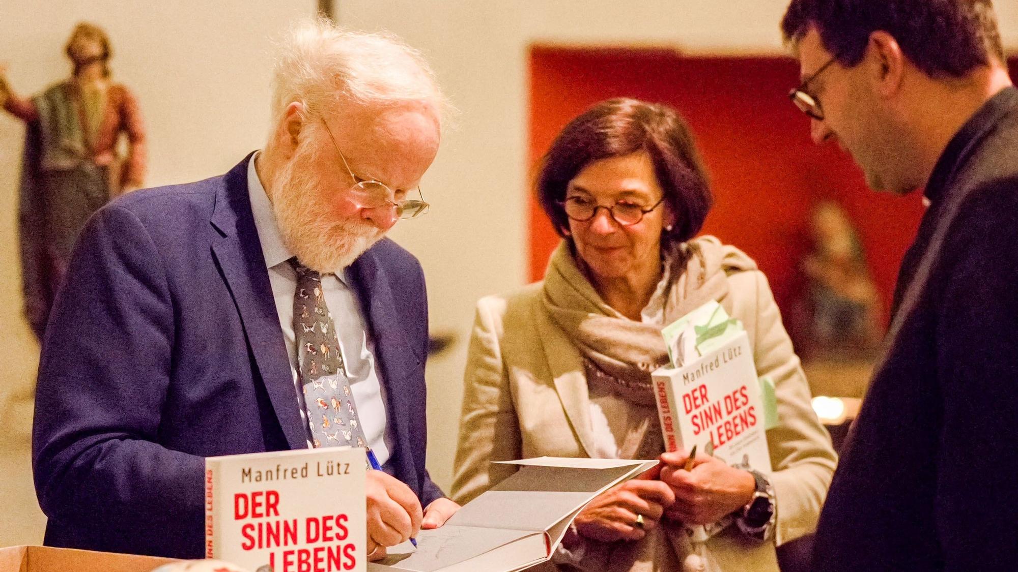 Manfred Lütz ist links im Bild, er signiert ein Buch. Neben ihm stehen eine Frau und ein Mann.