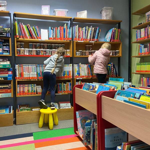 Zu sehen sind zwei Kinder, die vor einem großen Bücherregal stehen.