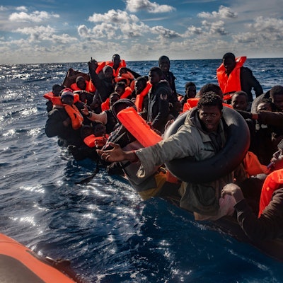 Mehrere Migranten sitzen in einem Boot im Mittelmeer, während Rettungskräfte versuchen ihnen zu helfen