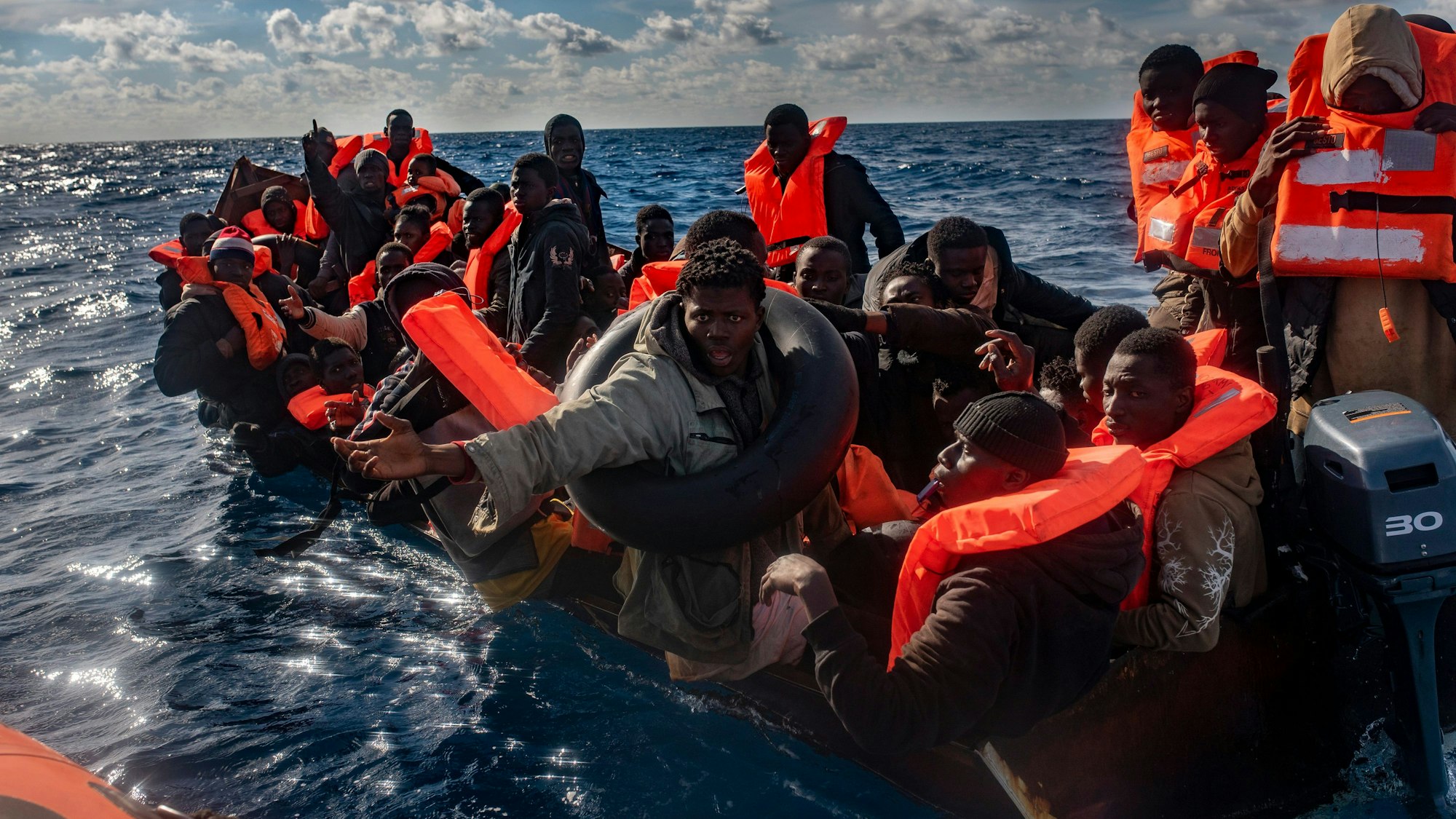 Mehrere Migranten sitzen in einem Boot im Mittelmeer, während Rettungskräfte versuchen ihnen zu helfen
