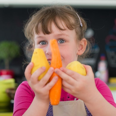 Ein lachendes Kind hält sich eine Möhre und Kartoffeln vors Gesicht.