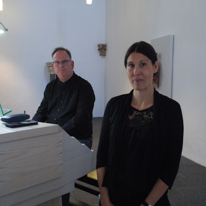 Eine Frau steht im Vordergrund. Hinter ihr sitzt ein Mann mit Brille.