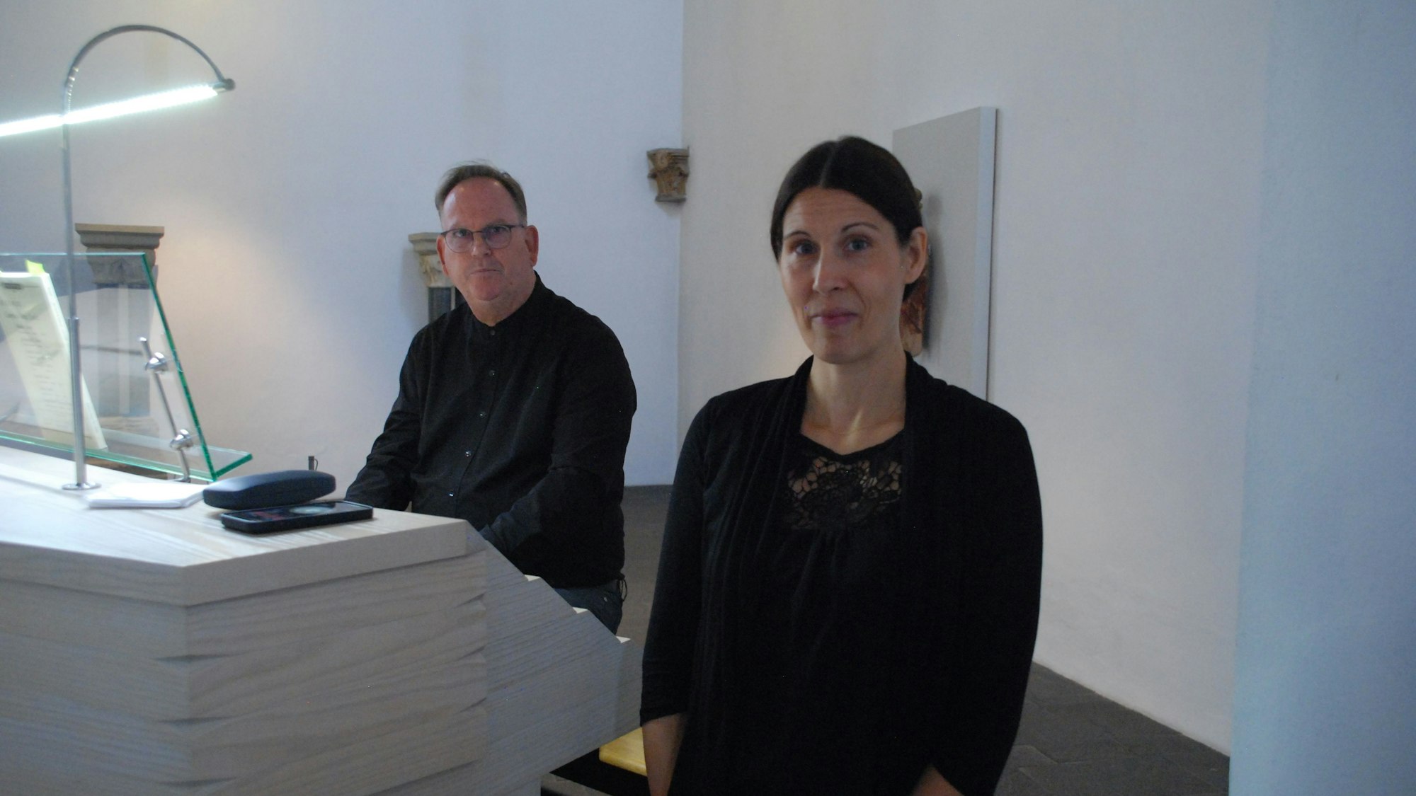 Eine Frau steht im Vordergrund. Hinter ihr sitzt ein Mann mit Brille.