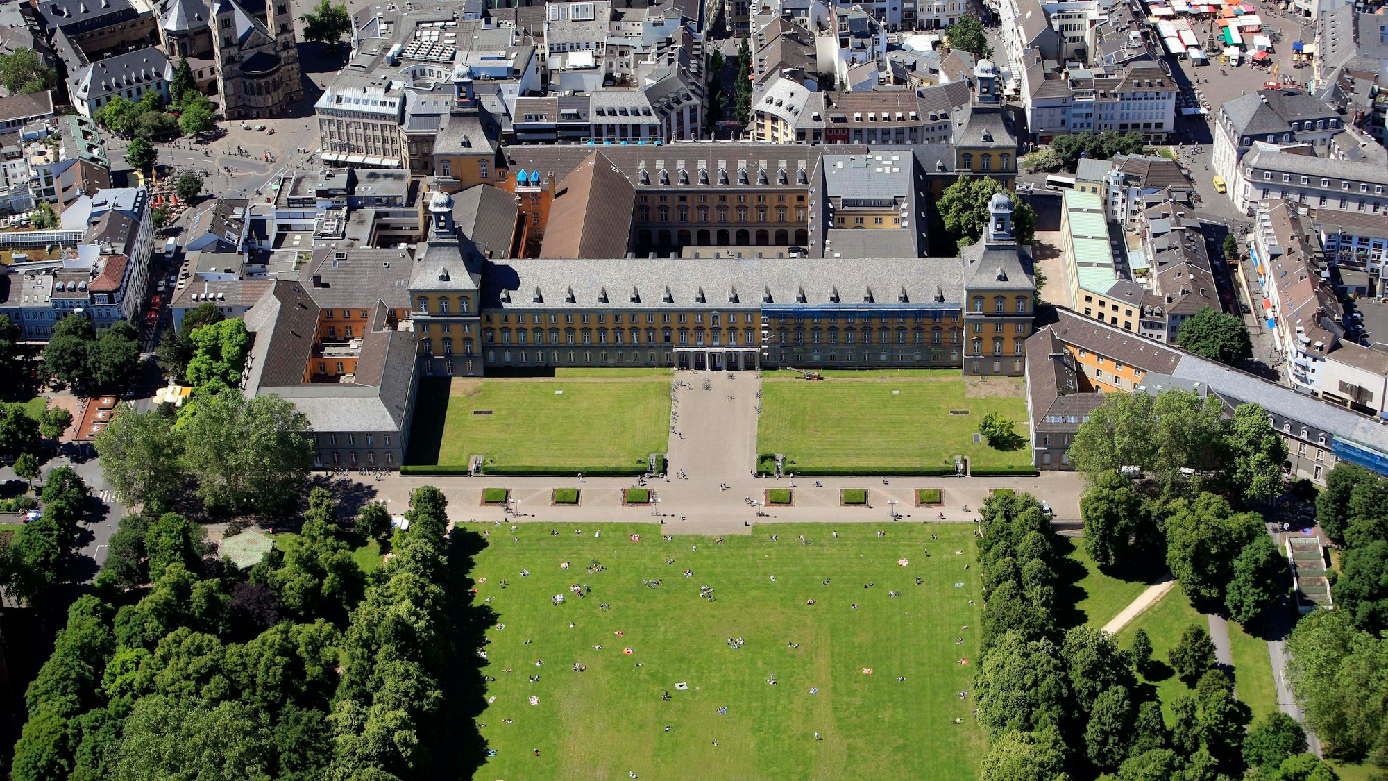 Das Hauptgebäude der Uni Bonn und der Hofgarten von oben. Einige Personen sitzen auf der Rasenfläche.