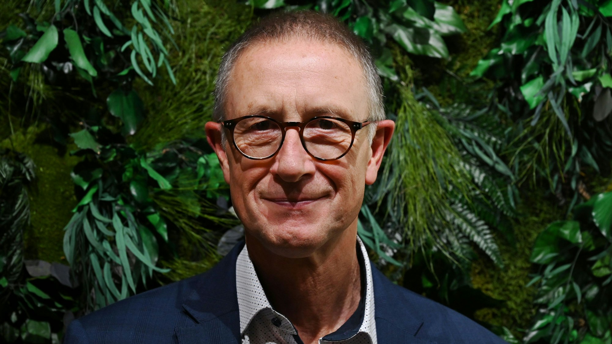 Helmut Schneider, Regionaldirektor der AOK Rheinland/Hamburg, steht vor einer Wand mit grünen Pflanzen.