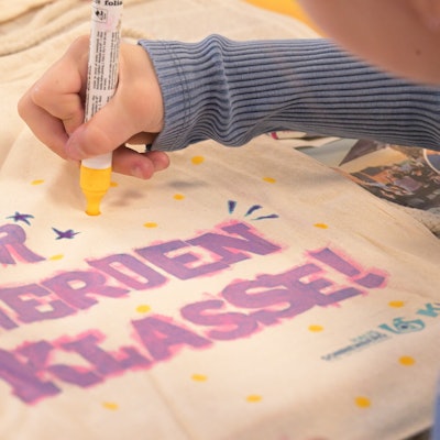 Ein Kind bemalt ein Stück Stoff mit dem Schriftzug: "Wir werden Klasse!"
