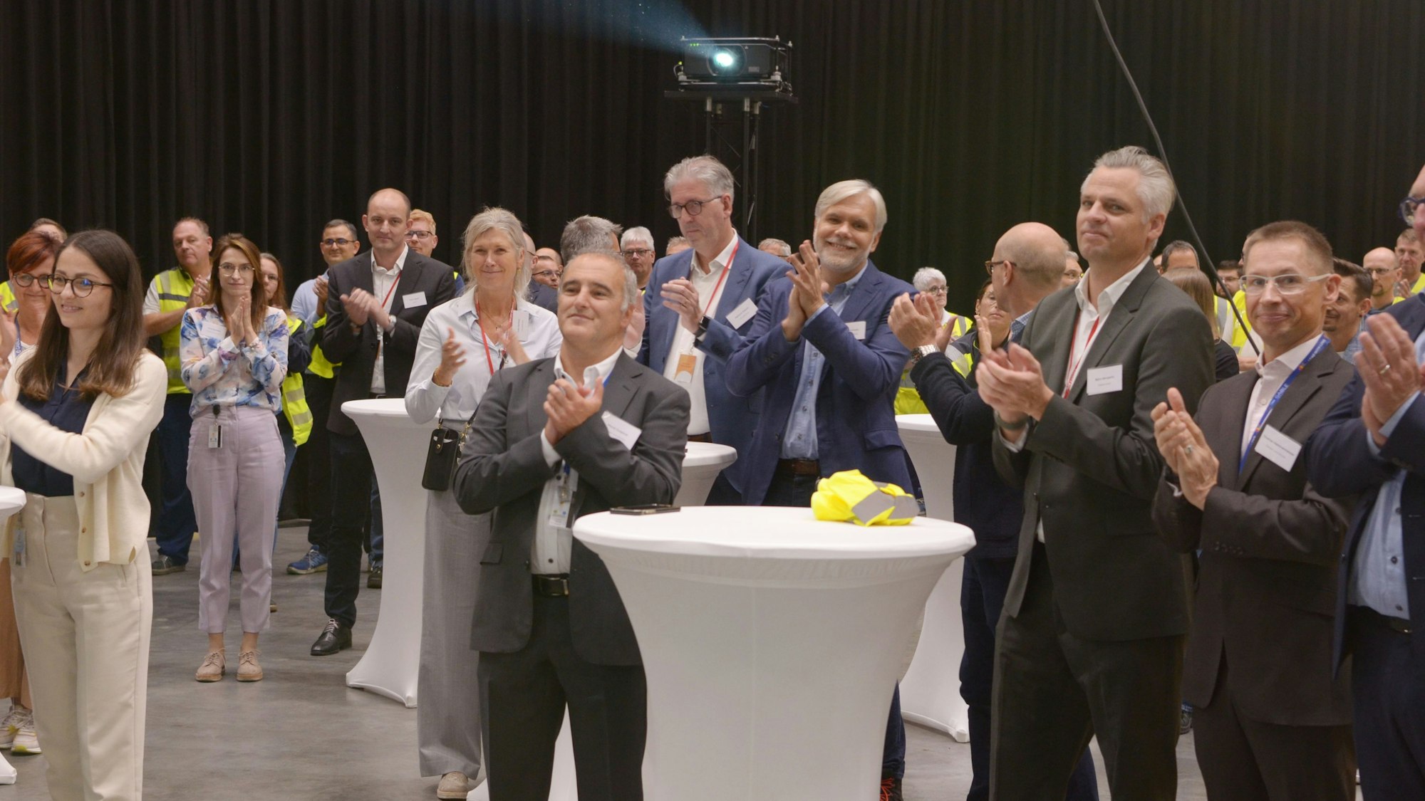 Die Gäste stehen applaudierend in einer Halle, wo die Eröffnungsreden gehalten werden.