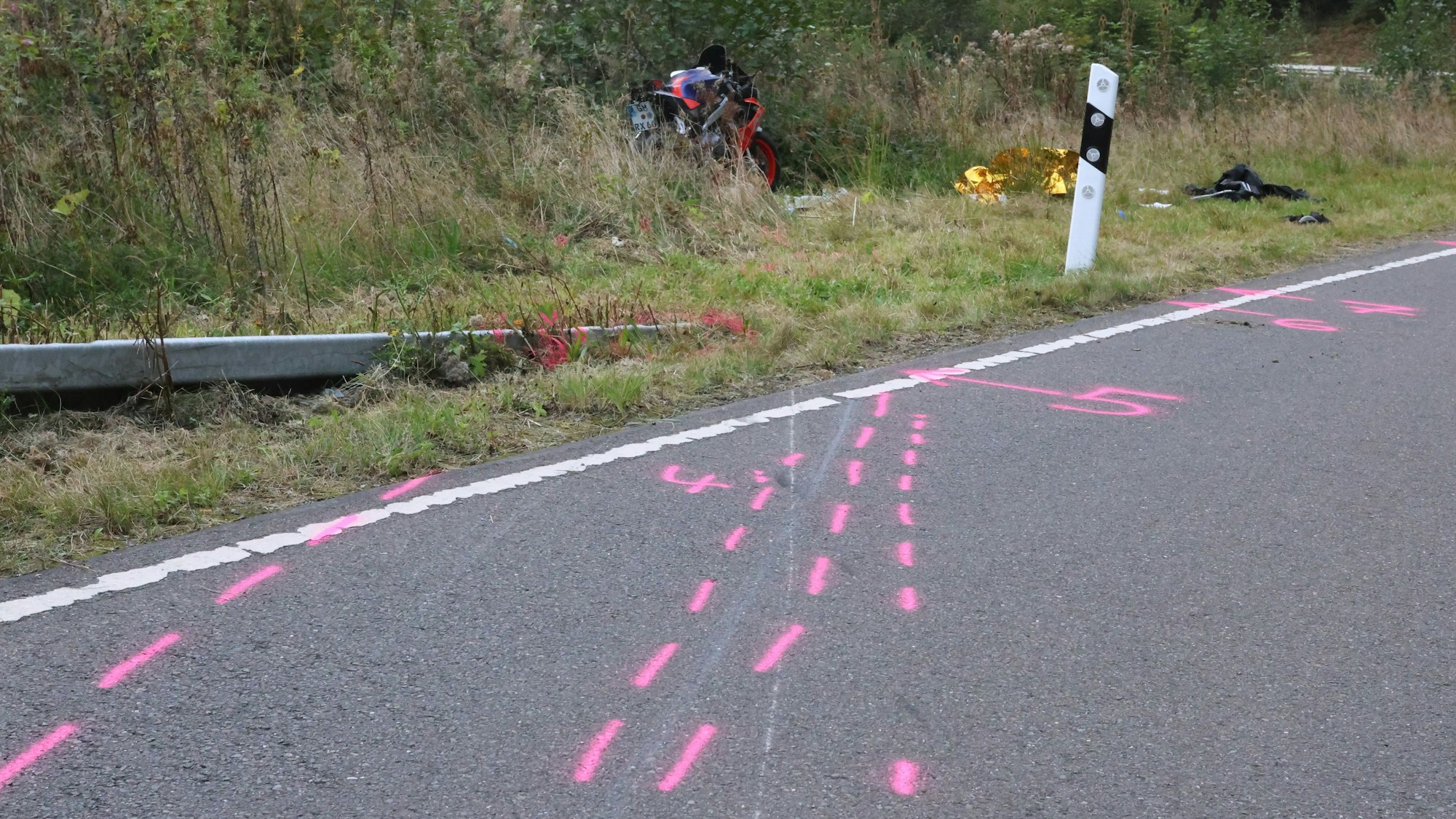 Auf einer Straße und dem abgesenkten Ende einer Leitplanke sind rosa Markierungen aufgesprüht. Ein verunfalltes Motorrad liegt in der Böschung.