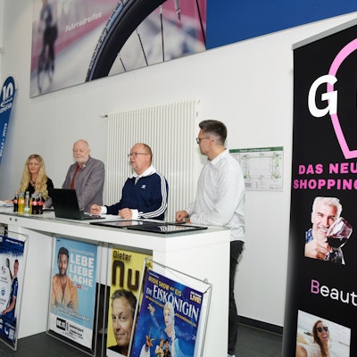 Menschen stehen an Tischen, mehrere Veranstaltungsplakate sind zu sehen.