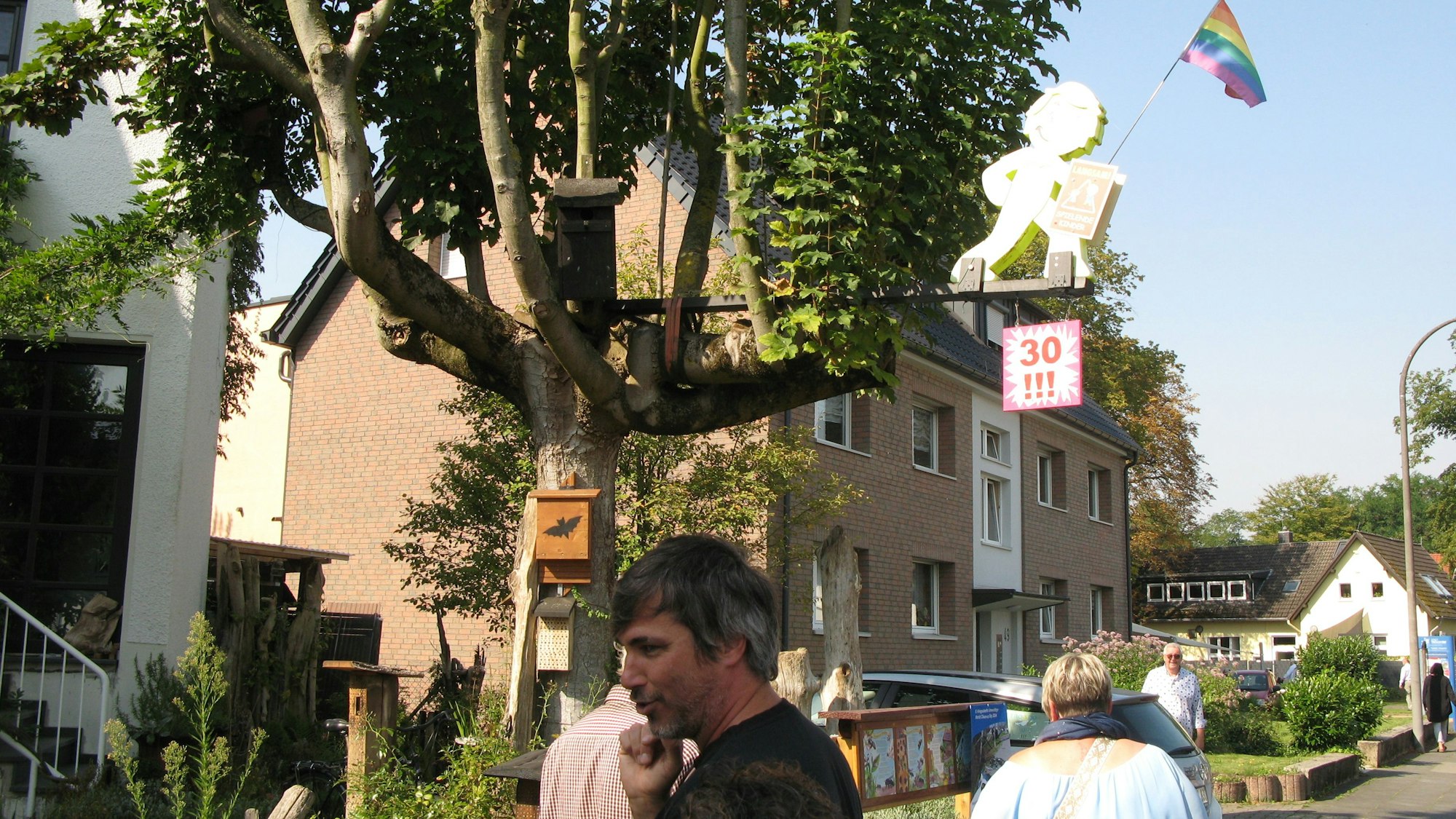 In einem Baum hängen ein Vogelnistkasten, ein Fledermauskasten, eine Kinderfigur mit Tempo-30-Schild und die Regenbogenflagge.