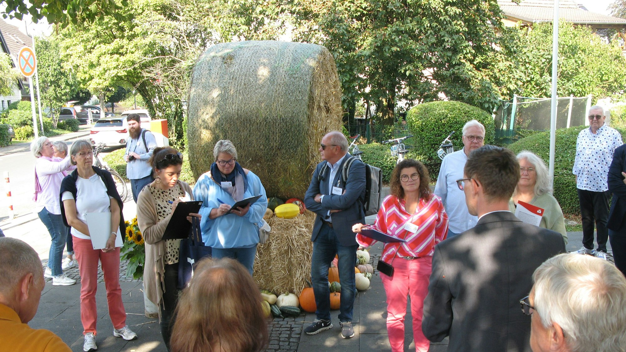 Männer und Frauen stehen auf einem Platz, der mit einer Strohballenfigur und Kürbissen geschmückt ist.