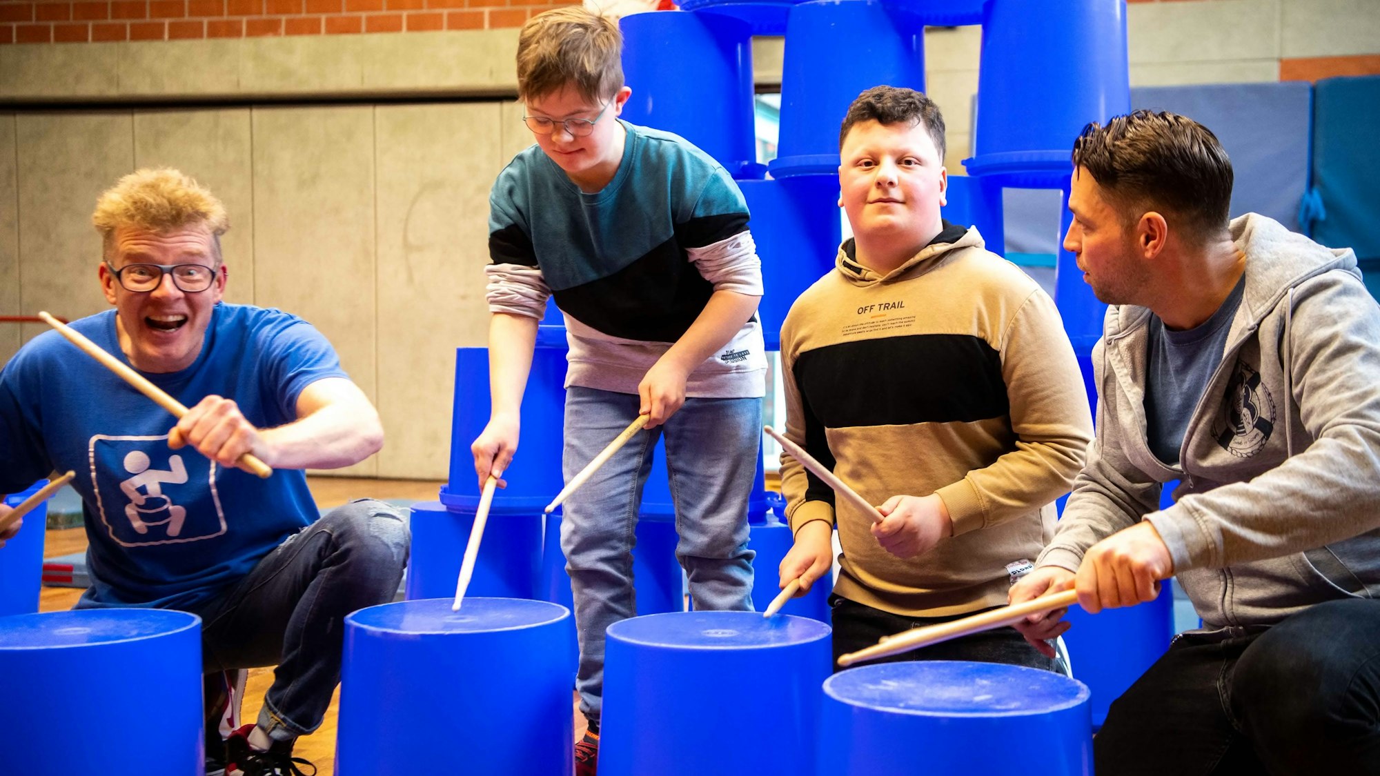 Das Bild zeigt zwei Schüler, die auf blaue Eimer eintrommeln. Zwei Lehrer unterstützen sie dabei.