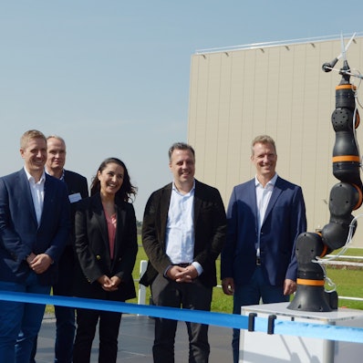 Vertreter des Unternehmens Procter & Gamble und Gäste stehen hinter einem blauen Band. Sie beobachten, wie ein Roboter eine Schere in Position bringt, die das Band durchtrennen soll.